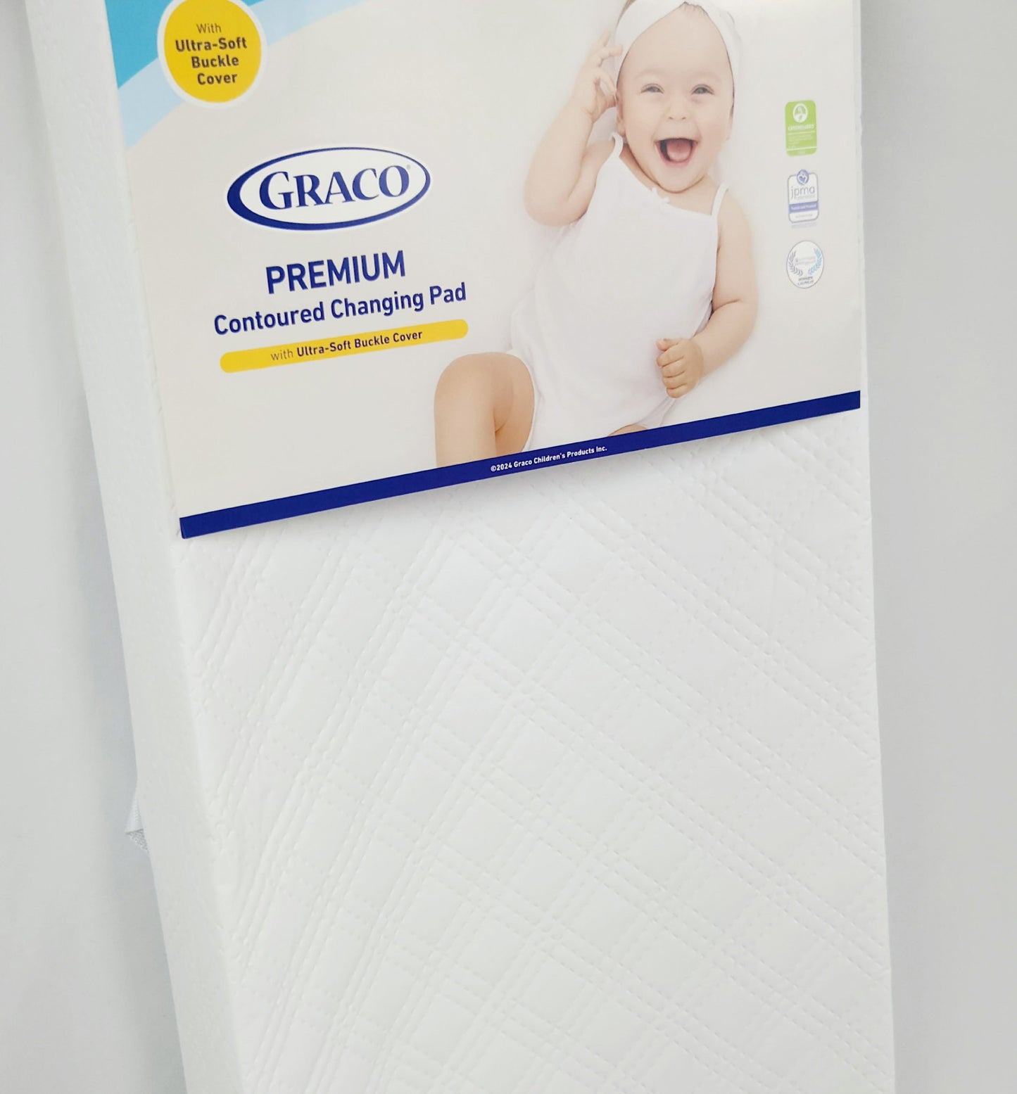 Graco Premium Contoured Changing Pad in white, with a visible blue and white packaging showing the product name and a baby image.